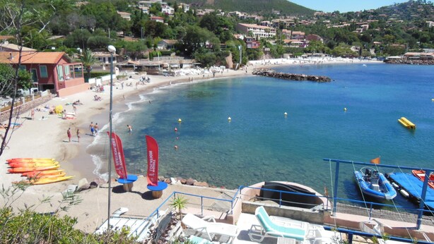Une plage à l’eau d’un bleu limpide, bordée d’un rivage sablonneux, avec en arrière-plan des maisons et des collines.