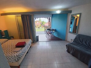 Une pièce comprenant un lit, un canapé, une table et une chaise. On peut voir, par la fenêtre, un balcon avec une balustrade et quelques plantes.