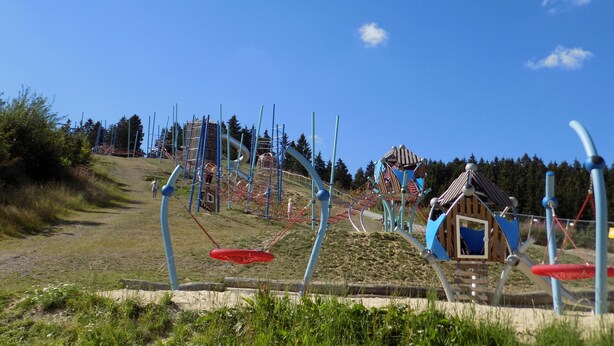 Un terrain de jeux doté de diverses installations, notamment des toboggans et des balançoires, aménagé sur une colline verdoyante sous un ciel bleu dégagé.