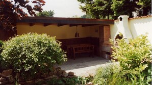 Un espace extérieur abrité, avec un banc et une table, entouré de verdure et d’un mur en pierre.