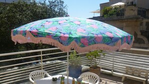 Un balcon avec une table blanche, des chaises, un parasol coloré, des plantes en pot et un banc.