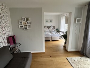 Uma sala de estar moderna com um sofá cinzento, uma mesa de centro e uma parede decorada com quadros. Uma cama com cabeceira cinza e roupa de cama branca é visível ao fundo.