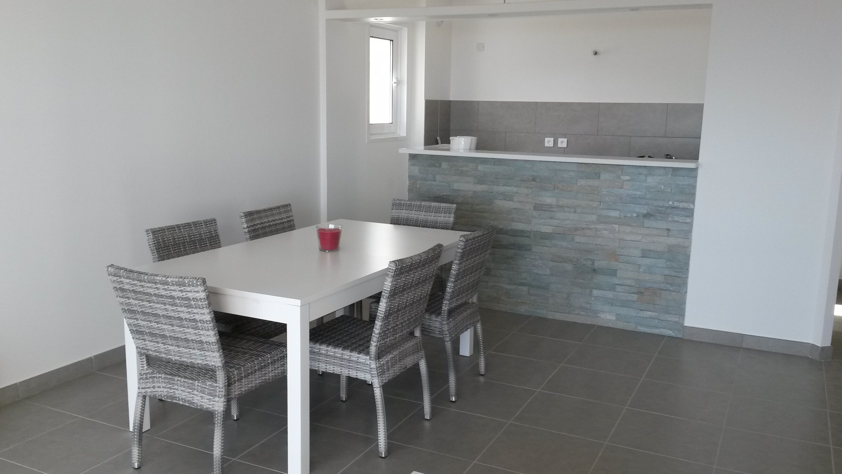 Un comedor con mesa blanca y sillas de mimbre, una cocina con azulejos grises y azules, y una ventana sobre el fregadero.