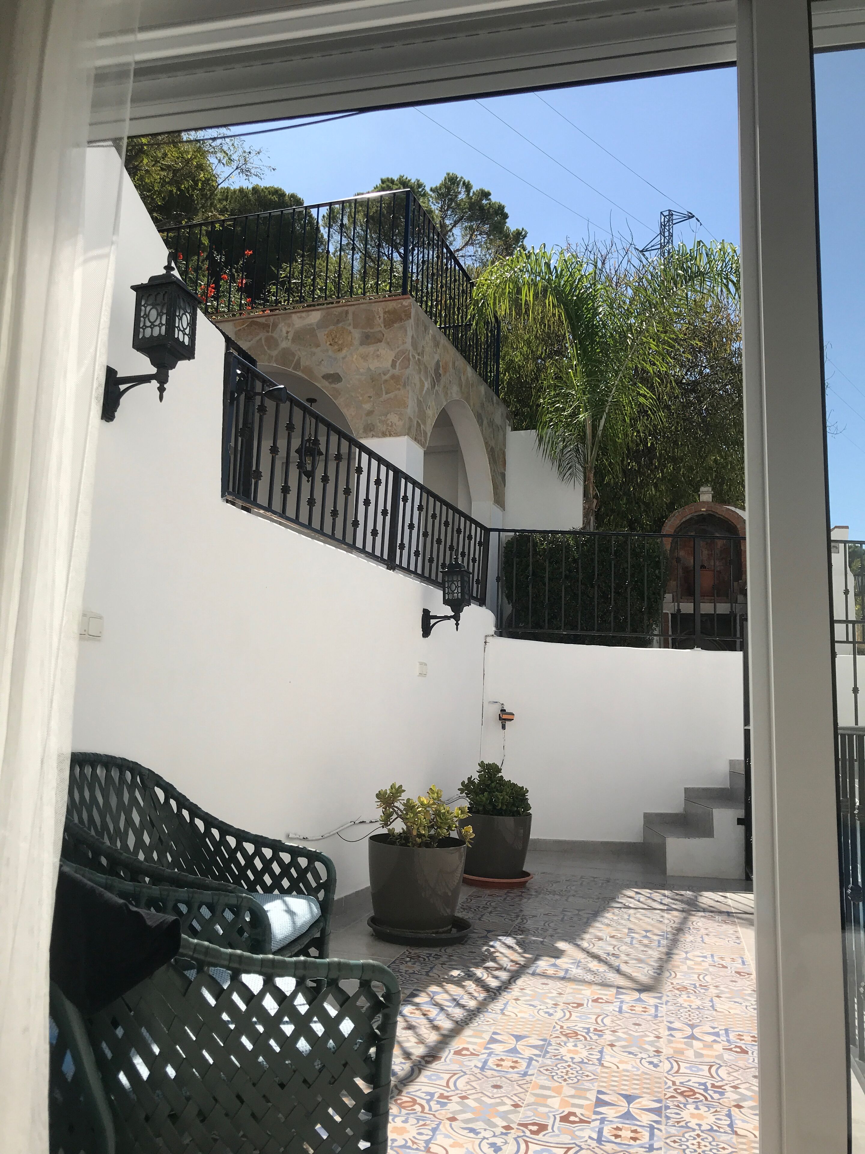 Une cour avec un mur blanc, un escalier en pierre, une rambarde en métal et deux plantes en pot.