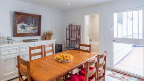 A dining area with a wooden table, chairs, and a bowl of fruit. There is a painting on the wall and a metal shelf with plants. - Typical white Andalusian house in the heart of PUEBLO, with swimming pool and jacuzzi (Alhaurín el Grande)
