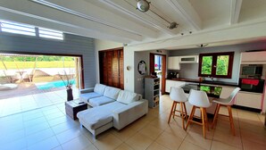 A modern living room with a sectional sofa, a dining area with white chairs, a kitchen with white cabinets, and a view of a swimming pool outside. - Unique in Guadeloupe: Villa 400 m from the beach with mango tree house (Le Gosier)