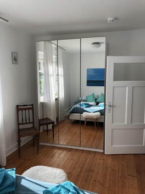 Un dormitorio con suelo de madera, cama con ropa de cama azul, una puerta blanca y un espejo.