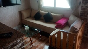 A living room with a sofa, a glass coffee table, a television mounted on the wall, a window with curtains, and a brick wall. - Studio next to Macdonald center agdal (Rabat)