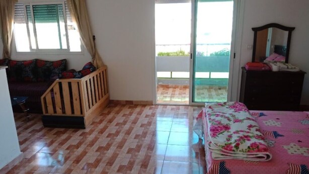A room with a wooden bed, a small wooden bench, a window with curtains, a sliding glass door, and a dresser with a mirror. - Studio next to Macdonald center agdal (Rabat)