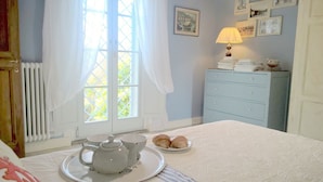 A bedroom with a bed, a dresser, a window with curtains, and a bedside table with a teapot, cups, and pastries.