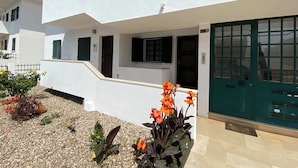 Ein weißes Gebäude mit einer grünen Tür, ein kleiner Garten mit orangefarbenen Blumen und eine geflieste Terrasse.