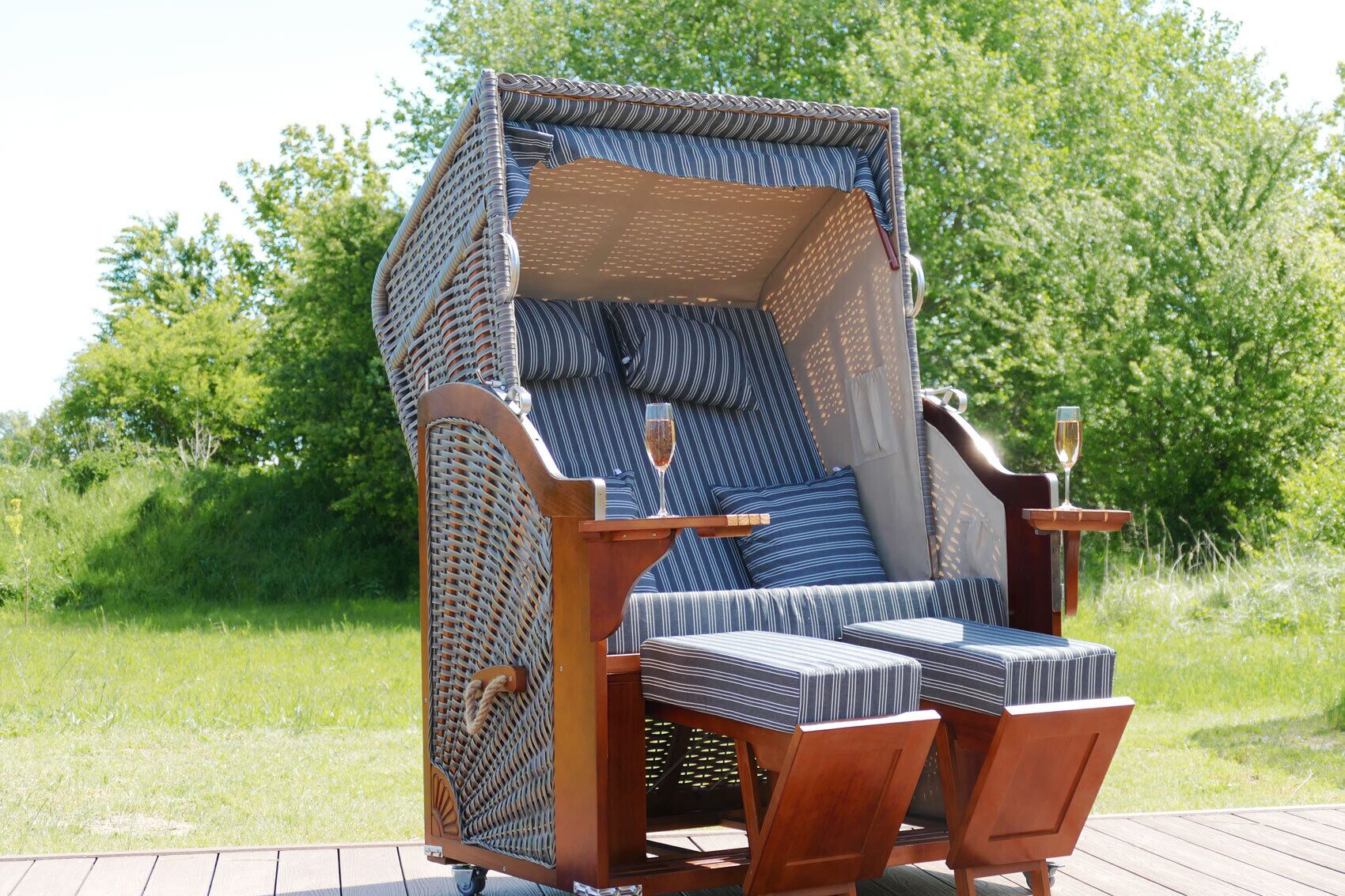 A wicker lounge chair with a cushioned seat and backrest, positioned on a wooden deck with a glass of champagne.