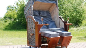 Een rieten lounge stoel met een kussenzit en rugleuning, geplaatst op een houten terras met een glas champagne.