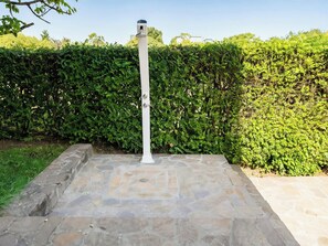 Une douche extérieure blanche, dotée d’un sol en pierre et entourée d’une haie.
