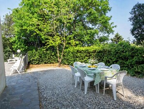 An outdoor dining area with a table set for four, surrounded by white plastic chairs, a gravel ground, and a backdrop of lush green trees and hedges. - panoramica casa con piscina privata (siena)