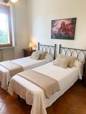A bedroom with two beds, a nightstand with a lamp, a window, and a painting on the wall.