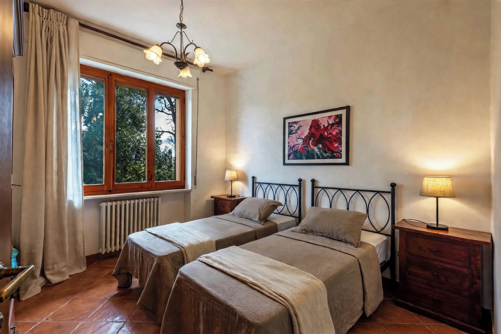 Une chambre avec deux lits, un radiateur, une fenêtre avec des rideaux, un lustre, un tableau et des lampes de chevet.