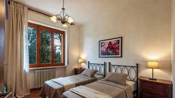 Une chambre avec deux lits, un radiateur, une fenêtre avec des rideaux, un lustre, un tableau et des lampes de chevet.