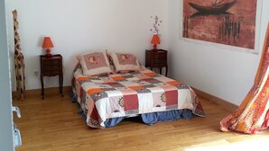 Een slaapkamer met een houten vloer, een bed met een kleurrijke quilt, twee nachtkastjes met lampen, een aan de muur hangend schilderij en een gordijn.