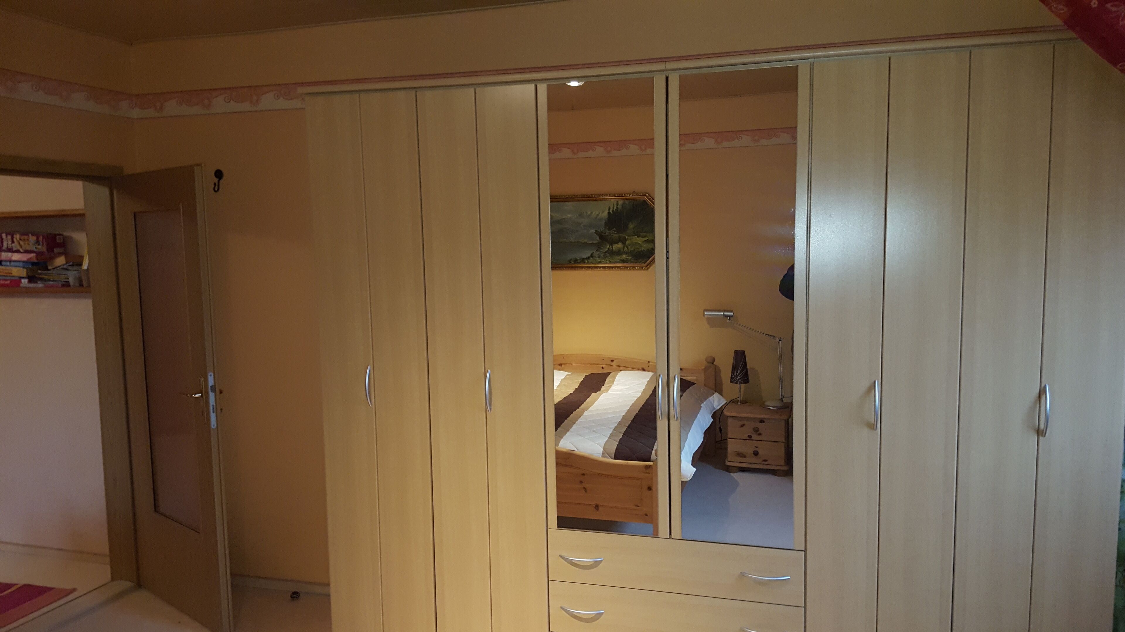 Een slaapkamer met een grote inbouwkast, een bed met een gestreept dekbedovertrek, een nachtkastje met een lamp, en een wandplank met boeken.