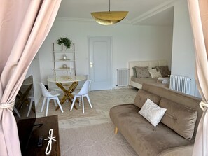 Een woonkamer met een bruine bank, witte stoelen, een ronde tafel, een witte deur en een radiator.