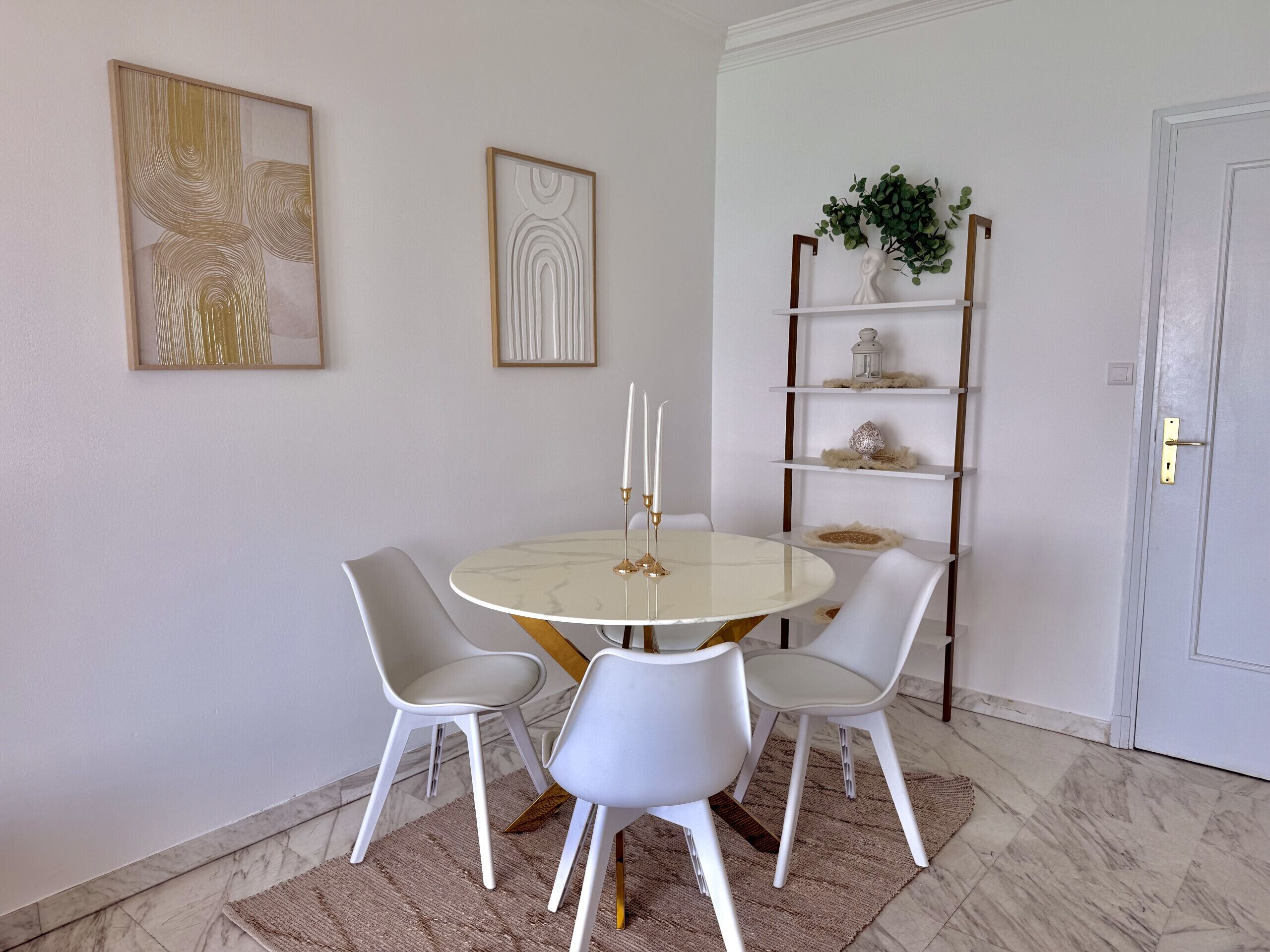 A dining area with a round table, four white chairs, two framed artworks, a plant on a shelf, and a white door.