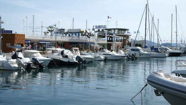 Een jachthaven met meerdere boten afgemeerd, een gebouw met de naam 'L'AMIRAL' en vlaggen, en een restaurant aan het water.