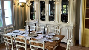 Comedor con una mesa de madera dispuesta para cuatro comensales, rodeada por sillas blancas con asientos trenzados. Un aparador blanco con puertas de cristal exhibe distintos platos y cristalería.
