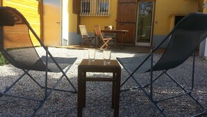 En utomhusplats med sittmöbler, bestående av två stolar och ett bord, med grusunderlag och en gul byggnad med dörr och fönster.