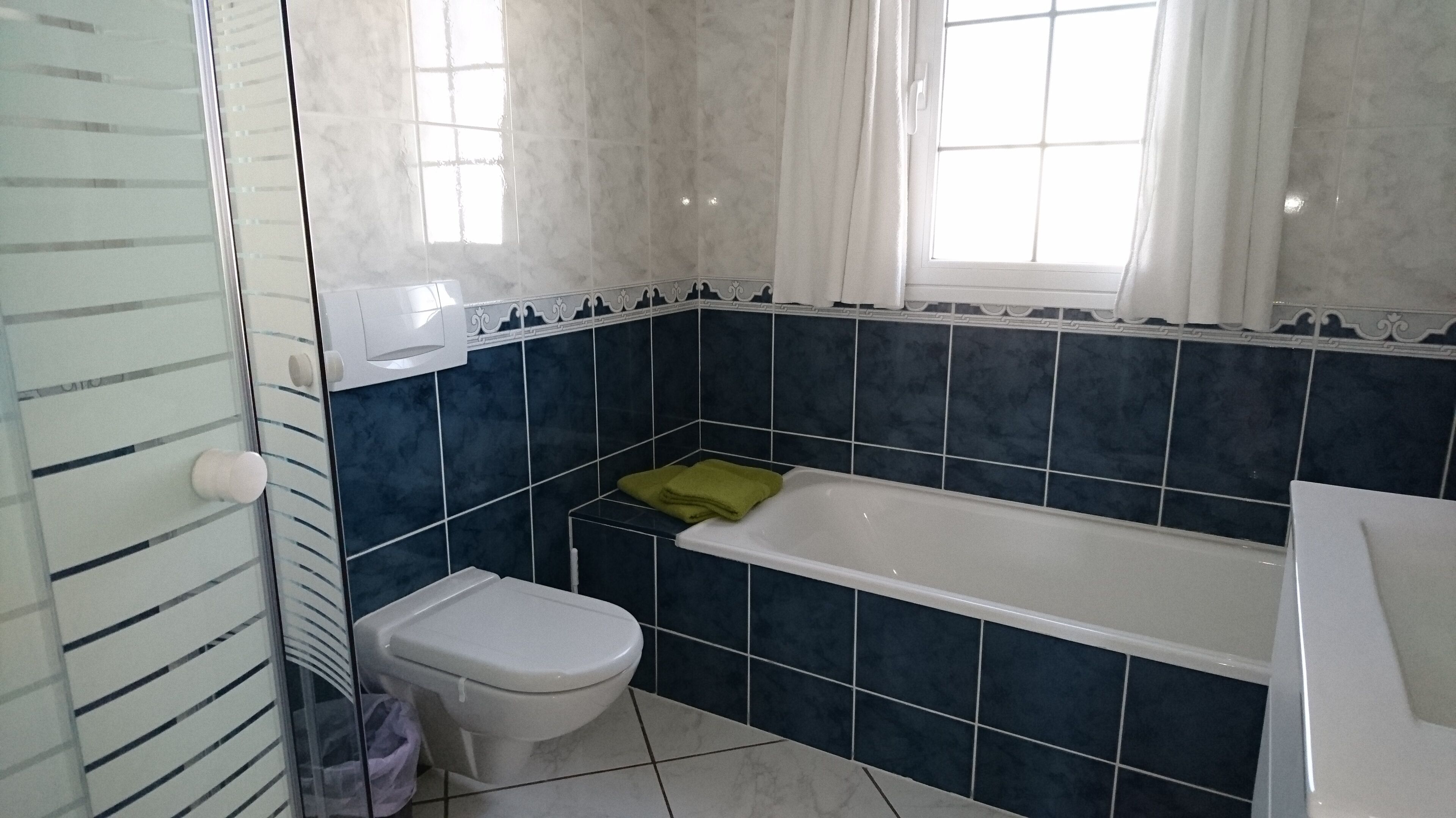Une salle de bain avec des toilettes blanches, une baignoire et une fenêtre avec des rideaux.