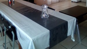 Un comedor con una mesa larga, sillas, un televisor montado en la pared, una lámpara de techo y una ventana con cortinas.