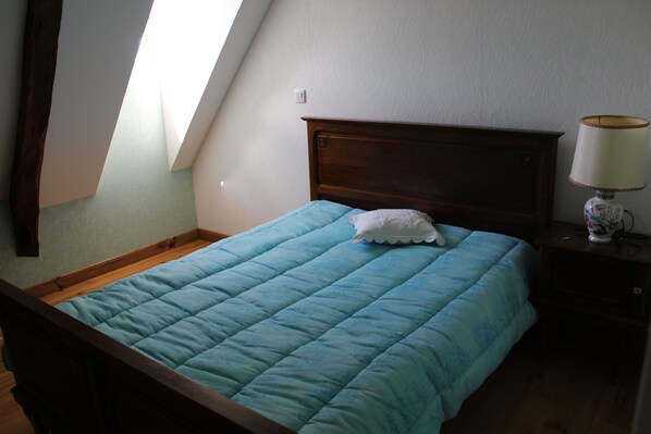 Una camera da letto con un letto con struttura in legno, una coperta trapuntata blu, un cuscino bianco, un comodino con una lampada e una finestra con trave in legno.