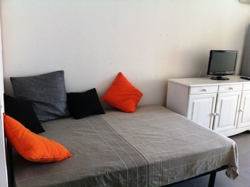 Un lit avec une couette grise, des oreillers noirs et orange, et une commode blanche sur laquelle se trouve un téléviseur.