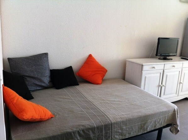 A bed with a grey quilt, black and orange pillows, and a white cabinet with a television. - Studio waterfront, near beach, sea view, 4 persons (Palavas-Les-Flots)