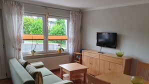 Ein Wohnzimmer mit einer Couch, einem Holztisch, einem an der Wand befestigten Fernseher und einem Fenster mit Vorhängen.