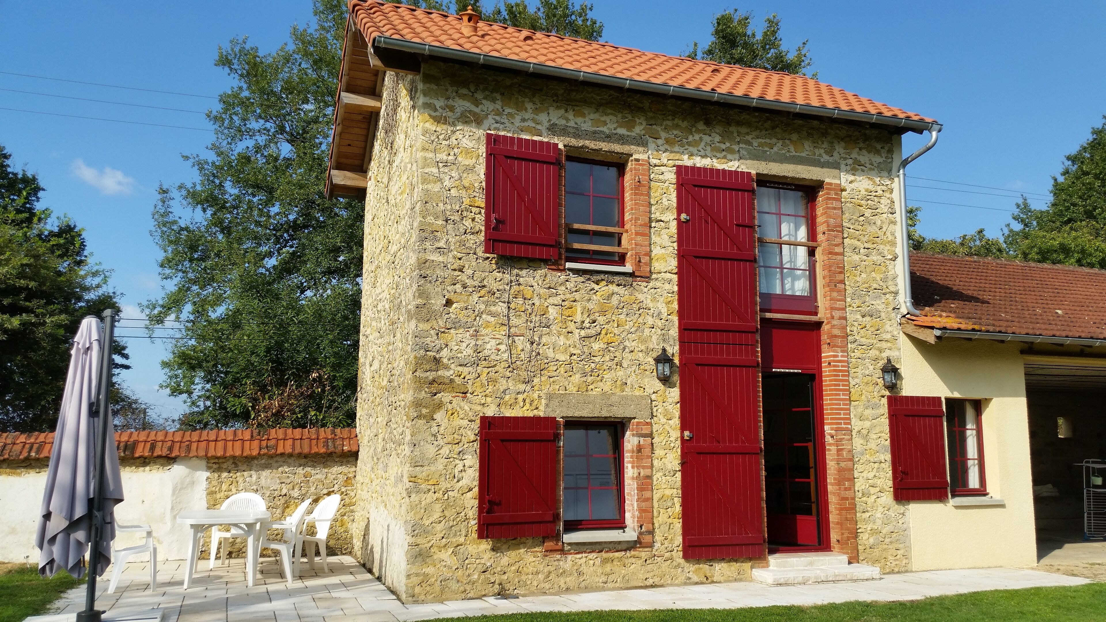 A two-story stone house with red shutters, a tiled roof, and a small patio with plastic chairs and a table.