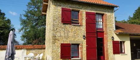A two-story stone house with red shutters, a tiled roof, and a small patio with plastic chairs and a table.