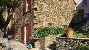 A stone building with red shutters, a small garden with potted plants, and a metal drying rack. - traditional stone village house (Olmeta-Di-Tuda)