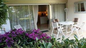 Une terrasse avec une table ronde et des chaises, une porte-fenêtre coulissante donnant sur une cuisine, et des fleurs de bougainvillier violettes au premier plan.