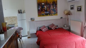 A bedroom with a red bedspread, a painting on the wall, and a kitchen area with a refrigerator and stove.