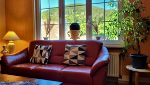 A red leather sofa with patterned cushions, a lamp, and a view of a green landscape through large windows.