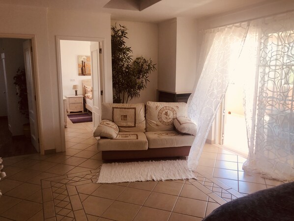 Una sala de estar con un sofá color beige, un tapete blanco y una planta grande. Se observa una puerta que da a otra habitación con una cama y una mesita de noche.