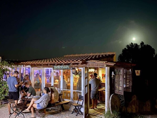 A small outdoor dining area with tables and chairs, a wooden structure with a tiled roof, and a sign that reads 'BOMBAR.'