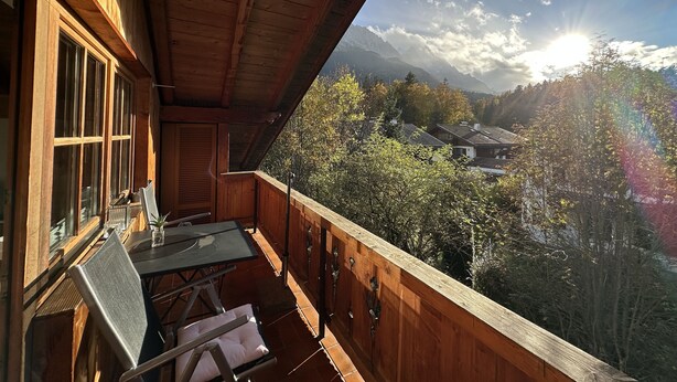 A wooden balcony with a table and chairs, overlooking a mountainous landscape with trees and a village.