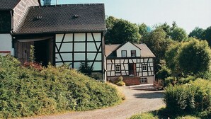 Une maison traditionnelle à colombages, avec un chemin de gravier menant à l’entrée, entourée de verdure.