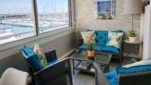 A room with a large window overlooking a marina, a blue sofa with floral cushions, a small table with a plant, and a lamp on a side table.