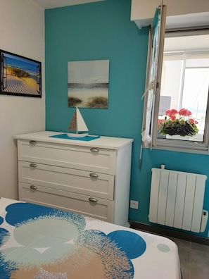 Une chambre avec un mur de couleur turquoise, une commode blanche, un lit recouvert d’une couvre-lit à motifs bleus et blancs, un tableau représentant une scène de plage, un petit modèle de voilier et une fenêtre donnant sur des fleurs et un bâtiment.