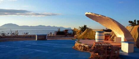 A pool area with a wooden deck, a large curved structure, and a view of the sea and mountains in the background.