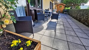 En udendørs terrasse med fletmøbler, et træbord og gule blomster i en plantekasse.
