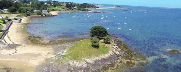 Vue aérienne d’une zone côtière avec une plage de sable, une petite île avec un arbre et de nombreux bateaux sur l’eau.